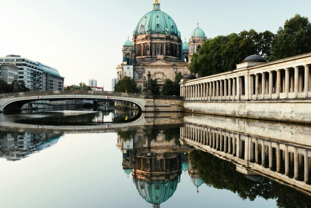Esta imagen presenta el icónico Berlín Dom, un impresionante edificio con cúpulas verdes, situado al lado de un canal de agua. Un puente cruza el agua, y la estructura se refleja perfectamente en la superficie del canal. Junto al lado hay edificios modernos y una hilera de columnas. Para profesionales en Perú que buscan conectarse con clientes B2B a través de LinkedIn, compartiendo contenido relacionado con los intercambios culturales y turísticos internacionales, mostrando atractivos como este, siguiendo las estrategias de "LinkedIn en Perú Cómo Conectar con Clientes B2B", pueden establecer relaciones valiosas y ampliar su red de negocios.