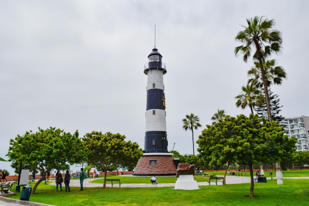Esta imagen presenta un majestuoso faro de colores blanco y azul - marino, ubicado en un parque verde rodeado de palmeras y árboles. Hay personas disfrutando del entorno, caminando y descansando en bancos. Para los creadores de contenido y propietarios de sitios web en Perú, promocionando imágenes de atractivos turísticos locales como este faro en Pinterest puede ser una estrategia efectiva. Siguiendo las prácticas de "Pinterest en Perú Cómo Generar Tráfico a Tu Web", se pueden captar la atención de usuarios interesados en viajar y conocer Perú, y así, llevar más tráfico a su sitio web.