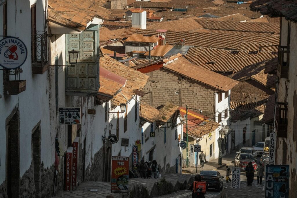 Esta imagen muestra una calle en una ciudad peruanas, con edificios de paredes blancas y techos de tejas rojas. Hay varios letreros publicitarios, algunos relacionados con tours y servicios turísticos, y personas caminando o en sus automóviles. Para aquellos en Perú que quieren ganar dinero a través de Twitch con transmisiones en vivo, mostrando la vida cotidiana y los atractivos turísticos locales de lugares como esta calle, siguiendo las estrategias de "Twitch en Perú Gana Dinero con Transmisiones en Vivo", pueden atraer a una audiencia y obtener ingresos a través de sus streams.