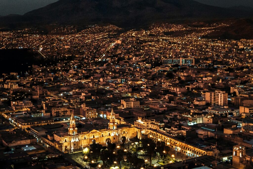 Esta imagen muestra una vista nocturna de una ciudad en Perú, iluminada por innumerables luces. En el centro destaca un importante edificio, probablemente un edificio histórico o un centro comunitario, con torres y una fachada bien iluminada. Las montañas en el fondo dan un contexto geográfico. Para los usuarios en Perú que deseen hacer que sus tweets tengan tendencia en Twitter, compartiendo contenido relacionado con la vida nocturna y los atractivos de ciudades peruanas, siguiendo las estrategias de "Twitter en Perú Cómo Hacer Tendencia Tus Tweets", pueden aumentar la interacción y la visibilidad de sus publicaciones.
